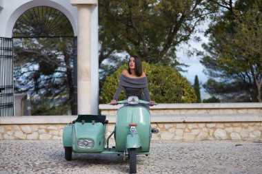 Genç, güzel, esmer, gri elbiseli İspanyol kadın sepetli antika bir motosiklet kullanıyor, dikkati dağınık görünüyor. Konsept motor, motosiklet, vintage, moda, güzellik.