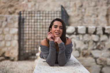 Genç, güzel, esmer, İspanyol kadın portresi, gri bir elbise giyiyor, gülümsüyor ve mutlu, elleri çenesinde, taş bir duvarda yatıyor. Kavram moda, güzellik, moda, kadınlık.