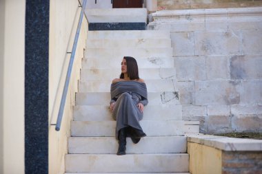 Genç, güzel, esmer, İspanyol kadın, gri elbiseli ve çizmeli, sakin, dikkati dağılmış bir şekilde merdivenlerde oturuyor. Kavram moda, güzellik, moda, kadınlık.