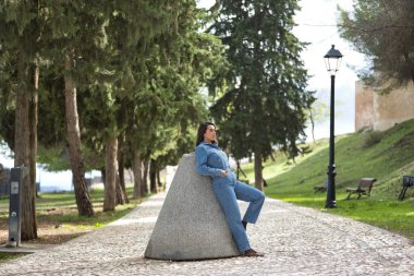 Genç, güzel, esmer, İspanyol bir kadın, kot elbise giyiyor, sonsuzluğa bakıyor, eski bir değirmen taşına yaslanıyor, taş bir yolda. Kavram moda, güzellik, moda, kadınlık.