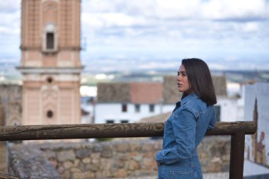 Genç, güzel, esmer, İspanyol kadın portresi, kot giyiyordu, utangaç ve çekingendi, arkası dönük, ahşap bir korkuluğa yaslanmıştı, arka planda kentsel bir manzara vardı..