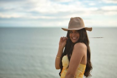 Genç, güzel, koyu renk saçlı, sarı elbiseli ve şapkalı bir kadının portresi, kameraya bakıp gülümseyen ve mutlu bir şekilde arka planda deniz olan bir çiviyle. Güzelliği, denizi kavra.