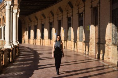 Genç bir kadın, güzel, esmer, siyah süslü elbiseli, gülümseyen, İspanya meydanında yürürken Sevilla 'da. Güzelliği, modayı, modayı, kadınlığı kavra.