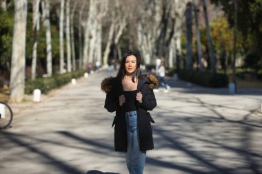 Genç, güzel, siyah saçlı, kürklü kapüşonlu bir kadın, siyah tişört ve kot pantolon, soğuk, güneşli bir günde ağaçlık bir sokakta yürüyor. Kış kavramı, soğuk, sonbahar, hava.