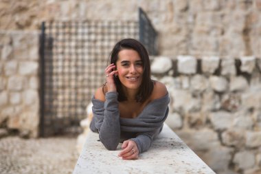 Genç, güzel, esmer, İspanyol bir kadının portresi, gri bir elbise giyiyor, saçlarına dokunuyor, gülümsüyor ve mutlu, taş bir duvarda yatıyor. Kavram moda, güzellik, moda, kadınlık.