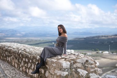 Genç, güzel, esmer, İspanyol kadın, gri bir elbise ve çizme giyiyor, rahat ve sakin, taş bir duvarda oturuyor, arkasında bulutlardan ve kırsal alanlardan oluşan güzel bir manzara var..