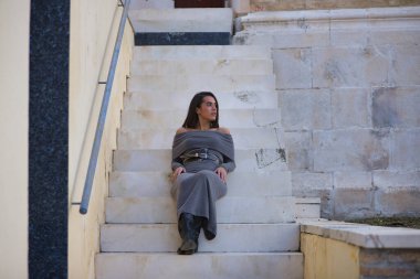 Genç, güzel, esmer, İspanyol kadın, gri elbiseli ve çizmeli, sakin, dikkati dağılmış bir şekilde merdivenlerde oturuyor. Kavram moda, güzellik, moda, kadınlık.