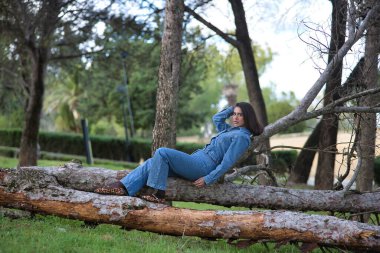 Genç, güzel, esmer, İspanyol bir kadın, kot elbise giyiyor, rahat ve sakin bir şekilde kameraya bakıyor, bir ağacın gövdesinde yatıyor, doğada. Kavram moda, güzellik, moda, kadınlık.