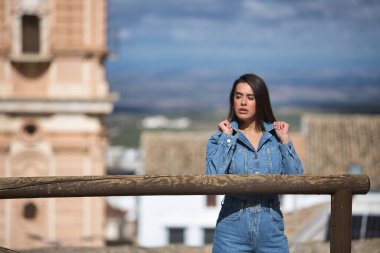 Genç, güzel, esmer, İspanyol bir kadın, kot elbise giyiyor, gömleğinin yakalarını kibirli bir tavırla kaldırıyor, arka planda kentsel bir manzara var. Kavram moda, güzellik, moda, kadınlık.