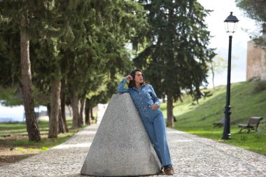 Genç, güzel, esmer, İspanyol bir kadın, kot elbise giyiyor, sonsuzluğa bakıyor, eski bir değirmen taşına yaslanıyor, taş bir yolda. Kavram moda, güzellik, moda, kadınlık.