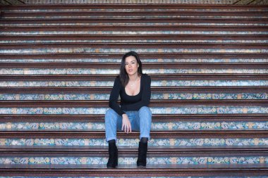 Genç, güzel, esmer, siyah uzun kollu tişört ve kot giyen, kameraya bakan, Seville 'de İspanya meydanında bir merdivende poz veren kadın. Güzellik, moda, kadınlık, turizm kavramları.