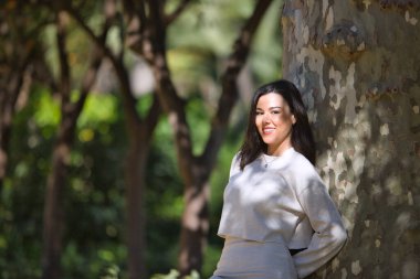 Yüzünde yaprakların gölgesi olan bir ağacın gövdesine yaslanmış beyaz elbiseli, genç, güzel, siyah saçlı bir kadın. Güzellik, moda, kadınlık, doğa, bitkiler, ağaçlar.