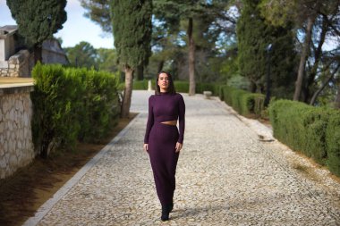 Genç, güzel, esmer, İspanyol bir kadın, mor bir elbise ve yüksek topuklu ayakkabı giyiyor, selvi ağaçlarıyla çevrili taş bir yolda tek başına yürüyor. Güzelliği, modayı, modayı kavra.