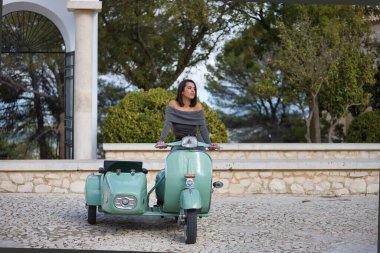 Genç, güzel, esmer, gri elbiseli İspanyol kadın sepetli antika bir motosiklet kullanıyor, dikkati dağınık görünüyor. Konsept motor, motosiklet, vintage, moda, güzellik.