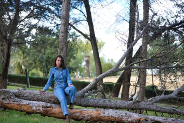 Genç, güzel, esmer, İspanyol bir kadın, kot elbise giyiyor, rahat ve sakin bir şekilde kameraya bakıyor, devrilmiş bir ağaç gövdesinde oturuyor, doğada. Kavram moda, güzellik, moda, kadınlık.
