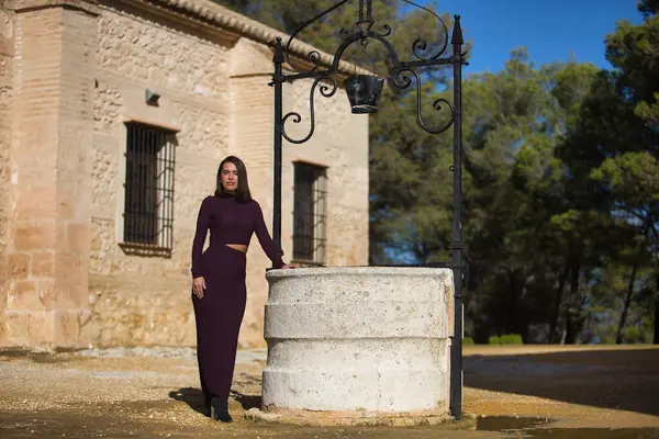Genç, güzel, esmer, İspanyol bir kadın, mor bir elbise ve topuklu ayakkabı giyiyor, kameraya bakıyor, eski bir su kuyusuna yaslanıyor. Güzelliği, modayı, modayı kavra.