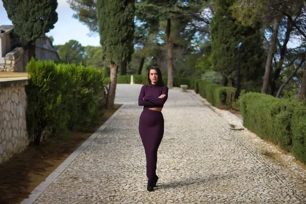 Genç, güzel, esmer, İspanyol bir kadın, mor bir elbise giyiyor, kolları çapraz, güçlü ve bağımsız, selvi ağaçlarıyla çevrili taş bir yolda. Güzelliği, modayı, modayı kavra.