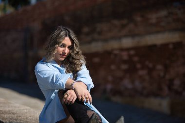 Genç, güzel, sarışın kadın, gömlek, deri etek ve botlar giyiyor, kameraya bakıyor, rahat ve sakin, taş bir duvarda oturuyor..