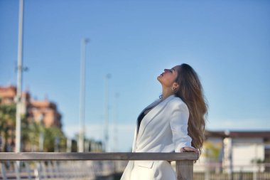 Genç bir kadın, güzel, kahverengi saçlı, beyaz ceketli, gözleri kapalı, saçı dökülmüş, rahat ve sakin, ahşap korkuluklara yaslanmış..