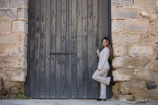 Genç, güzel, kahverengi saçlı, açık gri ceketli bir kadın, kameraya bakıyor, büyük, antika, ahşap bir kapının çerçevesine yaslanmış poz veriyor..