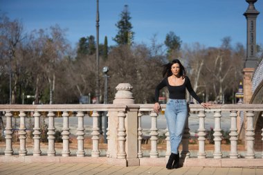 Genç, güzel, esmer bir kadın, siyah uzun kollu tişört ve kot pantolon giyiyor, saçları rüzgarda uçuşuyor, Seville 'de İspanya meydanında bir korkuluğa yaslanmış poz veriyor. Güzellik kavramı, kadınlık.