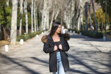 Genç, güzel, siyah saçlı, kürklü kapüşonlu bir kadın, siyah tişört ve kot pantolon, soğuk, güneşli bir günde ağaçlık bir sokakta yürüyor. Kış kavramı, soğuk, sonbahar, hava.