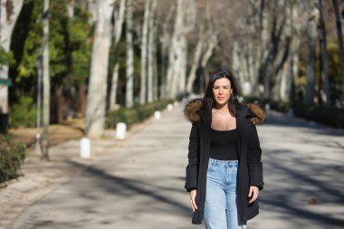 Genç, güzel, siyah saçlı, kürklü kapüşonlu bir kadın, siyah tişört ve kot pantolon, soğuk, güneşli bir günde ağaçlık bir sokakta yürüyor. Kış kavramı, soğuk, sonbahar, hava.