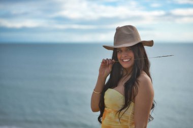 Genç, güzel, koyu renk saçlı, sarı elbiseli ve şapkalı bir kadının portresi, kameraya bakıp gülümseyen ve mutlu bir şekilde arka planda deniz olan bir çiviyle. Güzelliği, denizi kavra.