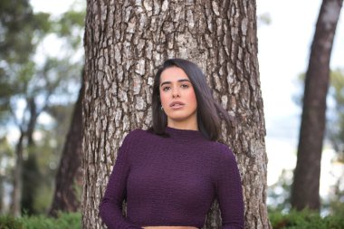 Mor elbiseli, genç, güzel, koyu saçlı, İspanyol bir kadının portresi, kameraya bakıyor, tatlı ve narin, büyük bir ağacın gövdesine yaslanmış. Güzelliği, modayı, eğilimi kavra.