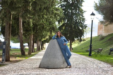 Genç, güzel, esmer, İspanyol bir kadın, kot elbise giyiyor, gülümsüyor ve mutlu görünüyor, taş bir yolda eski bir değirmen taşına yaslanıyor. Kavram moda, güzellik, moda, kadınlık.