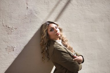 Genç, güzel, sarışın bir kadının portresi, yeşil bir ceket giyiyor, kolları çapraz, kameraya bakıyor, bağımsız ve güçlü, duvara yaslanmış..