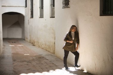 Genç, güzel, sarışın bir kadın, yeşil bir ceket, pantolon ve bot giyiyor, kameraya bakıyor, duvara yaslanıyor, ışığın ve gölgenin oynadığı ıssız bir sokakta.