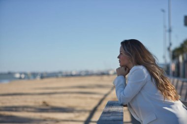 Genç, güzel bir kadın, kahverengi saçlı, beyaz ceketli, elleri çenesinde, ahşap korkuluklara yaslanmış sonsuzluğa bakıyor..