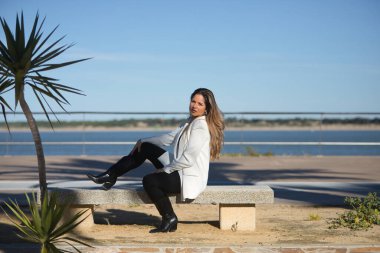 Genç, güzel, kahverengi saçlı, beyaz ceketli bir kadın, kameraya bakıyor, bir bankta yan oturuyor, yalnız ve sessiz, şafak vakti güneş ışınlarını alıyor..