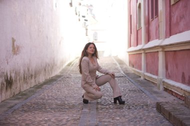 Açık gri takım elbiseli, genç, güzel, kahverengi saçlı bir kadın, çömelmiş, rahat ve sakin, bir köyde yalnız, Akdeniz tarzı bir sokakta poz veriyor..