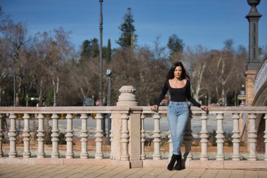 Genç, güzel, esmer bir kadın, siyah uzun kollu tişört ve kot pantolon giyiyor, saçları rüzgarda uçuşuyor, Seville 'de İspanya meydanında bir korkuluğa yaslanmış poz veriyor. Güzellik kavramı, kadınlık.