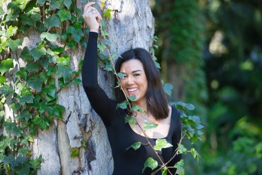 Genç, güzel, esmer bir kadın, siyah tişört ve kot pantolon, bir ağacın gövdesine yaslanmış, sarmaşıkla komik oynuyor, doğayla bağlantılı. Güzellik kavramı, kadınlık, doğa, bitkiler, ağaçlar.