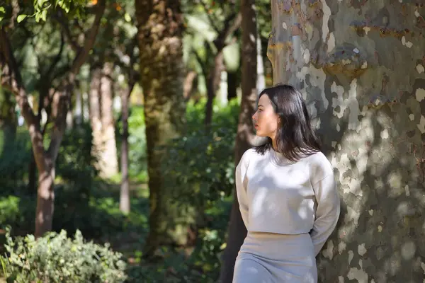 Yüzünde yaprakların gölgesi olan bir ağacın gövdesine yaslanmış beyaz elbiseli, genç, güzel, siyah saçlı bir kadın. Güzellik, moda, kadınlık, doğa, bitkiler, ağaçlar.