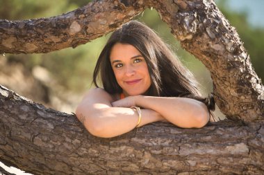 Turuncu elbiseli, genç, güzel, koyu renk saçlı bir kadının portresi, yumuşak ve gülümseyen, kumda oturan, devrilmiş bir ağacın gövdesine bakan. Güzelliği, mutluluğu, şefkati, sakinliği kavra..