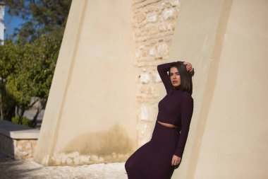 Genç, güzel, esmer, İspanyol bir kadın, mor bir elbise ve çizme giyiyor, duvara yaslanıyor, kameraya bakıyor, güneş ışınlarını alıyor. Güzelliği, modayı, modayı, yalnızlığı kavra.