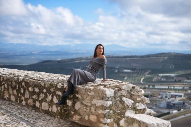 Genç, güzel, esmer, İspanyol kadın, gri bir elbise ve çizme giyiyor, sonsuzluğa bakıyor, taş bir duvarda oturuyor, arkasında bulutlardan ve kırsal alanlardan oluşan güzel bir manzara var..