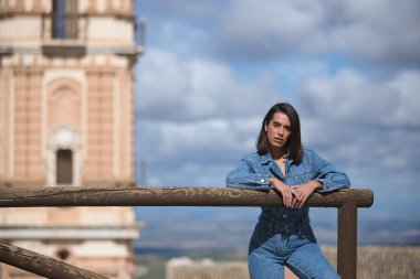 Genç, güzel, esmer, İspanyol bir kadın, rahat ve sakin bir şekilde kameraya bakıyor, arka planda şehir manzarası olan ahşap bir korkuluğa yaslanıyor..