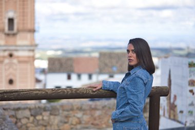 Genç, güzel, esmer, İspanyol kadın portresi, kot giyiyordu, utangaç ve çekingendi, arkası dönük, ahşap bir korkuluğa yaslanmıştı, arka planda kentsel bir manzara vardı..