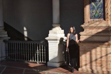 Genç bir kadın, güzel, esmer, siyah bir fantezi elbisesi, kayıp bir bakışla, tuğla bir duvara yaslanmış, ışık ve gölge oyunu oynuyor. Güzelliği, modayı, modayı, kadınlığı kavra.