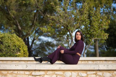 Genç, güzel, esmer, İspanyol bir kadın, mor elbiseli ve topuklu ayakkabılı, dikkati dağılmış bir bakışla duvarda oturuyor. Güzelliği, modayı, modayı kavra.