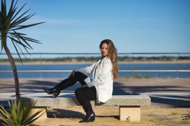 Genç, güzel, kahverengi saçlı, beyaz ceketli bir kadın, kameraya bakıyor, bir bankta yan oturuyor, yalnız ve sessiz, şafak vakti güneş ışınlarını alıyor..