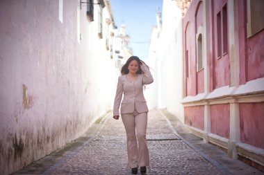 Genç, güzel, kahverengi saçlı bir kadın, açık gri bir ceket giyiyor, bir eli saçında, bir köyde yalnız, Akdeniz tarzı bir sokakta yürüyor..