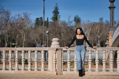 Genç, güzel, siyah uzun kollu tişört ve kot giyen esmer kadın, sonsuzluğa bakıyor, Seville 'de İspanya meydanında bir korkuluğa yaslanmış poz veriyor. Güzelliği, modayı, modayı, kadınlığı kavra