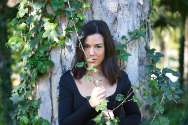 Siyah tişört ve kot pantolon giymiş, bir ağacın gövdesine yaslanmış ve doğayla bağlantılı, sarmaşığa dolanmış genç, güzel, koyu renk saçlı bir kadın. Güzellik kavramı, kadınlık, doğa, bitkiler, ağaçlar.