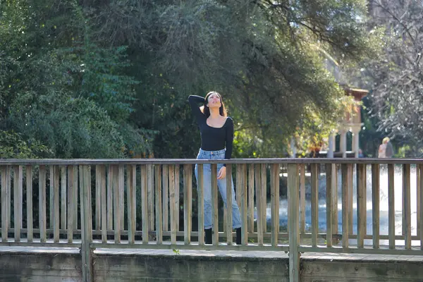 Siyah tişört ve kot pantolon giymiş, genç, güzel, esmer bir kadın. Tahta bir köprüde fotoğraf çekilirken poz veriyor. Güzelliği, modayı, eğilimi, kadınlığı kavra.